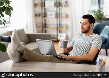technology, remote job and lifestyle concept - man with laptop computer drinking coffee and resting feet on table at home office. man with laptop drinking coffee at home