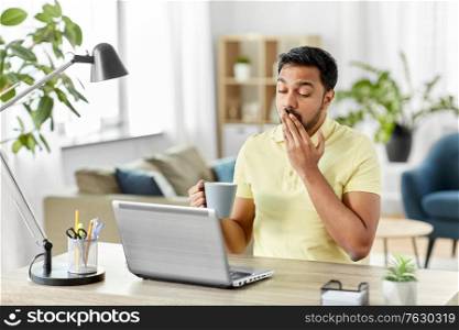 technology, remote job and lifestyle concept - sleepy tired indian man with laptop computer and coffee or tea yawning at home office. man with laptop and coffee yawning at home office