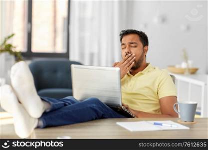 technology, remote job and lifestyle concept - tired yawning indian man with laptop computer resting feet on table at home office. tired man with laptop and feet on table at home