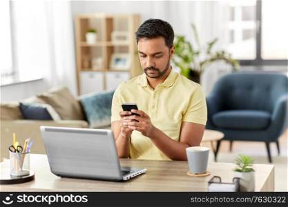 technology, remote job and people concept - indian man with smartphone and laptop computer at home office. happy indian man with smartphone at home office