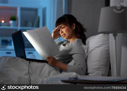 technology, remote job and people concept - stressed young asian woman with laptop computer and papers working in bed at home at night. stressed woman with papers working in bed at night
