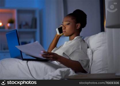 technology, remote job and people concept - young african american woman with laptop computer and papers calling on smartphone and working in bed at home at night. woman with laptop calling on smartphone in bed