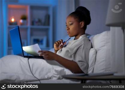 technology, remote job and people concept - young african american woman with laptop computer and papers working in bed at home at night. woman with laptop working in bed at night