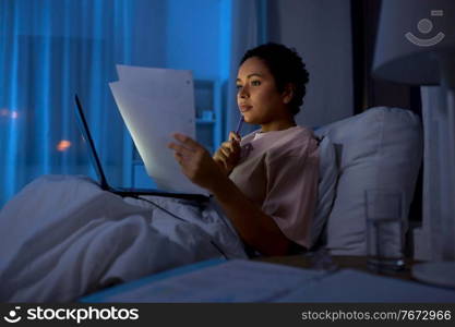 technology, remote job and people concept - young african american woman with laptop computer and papers working in bed at home at night. woman with laptop and papers in bed at night