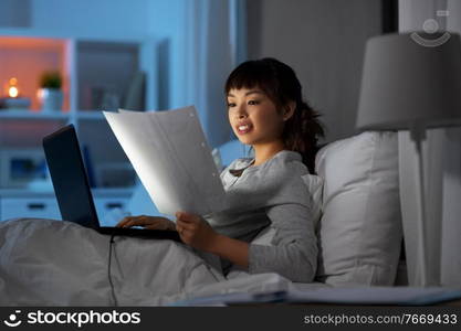 technology, remote job and people concept - young asian woman with laptop computer and papers working in bed at home at night. woman with laptop working in bed at night