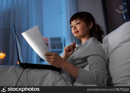 technology, remote job and people concept - young asian woman with laptop computer and papers working in bed at home at night. asian woman with laptop and papers in bed at night