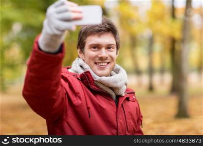 technology, season and people concept - happy smiling young man taking selfie by smartphone in autumn park