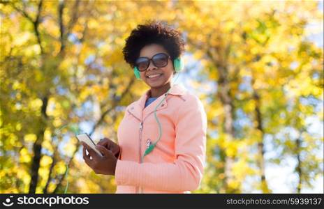 technology, season and people concept - smiling african american young woman or teenage girl with smartphone and headphones listening to music over autumn park background