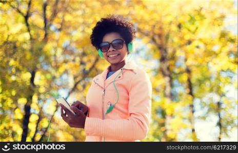 technology, season and people concept - smiling african american young woman or teenage girl with smartphone and headphones listening to music over autumn park background
