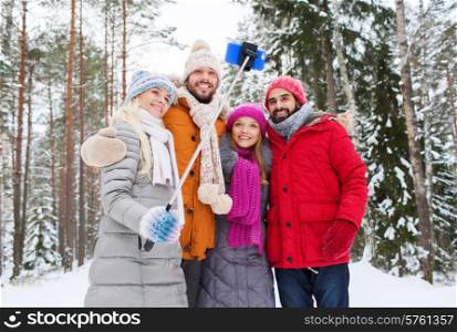 technology, season, friendship and people concept - group of smiling men and women taking selfie with smartphone and monopod in winter forest