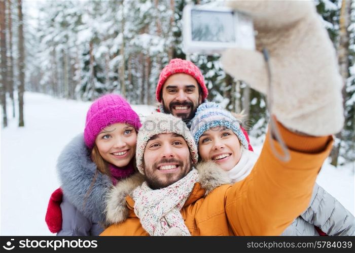 technology, season, friendship and people concept - group of smiling men and women taking selfie with digital camera in winter forest