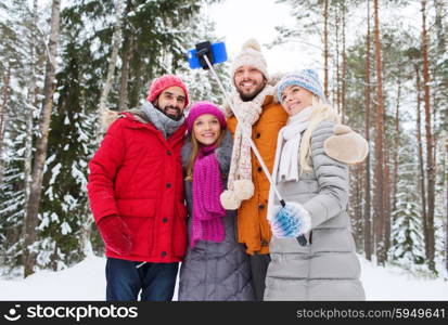 technology, season, friendship and people concept - group of smiling men and women taking selfie with smartphone and monopod in winter forest