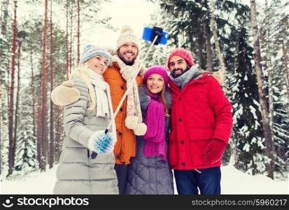 technology, season, friendship and people concept - group of smiling men and women taking selfie with smartphone and monopod in winter forest