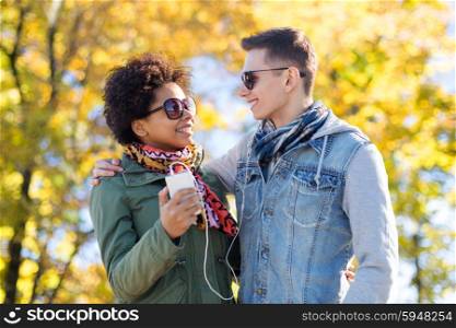 technology, season, people and friendship concept - smiling couple with smartphone and earphones listening to music over autumn park background