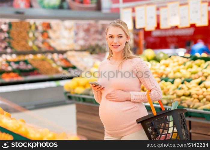 technology, shopping, food, pregnancy and people concept - happy pregnant woman with basket and smartphone at grocery store or supermarket. pregnant woman with shopping basket and smartphone