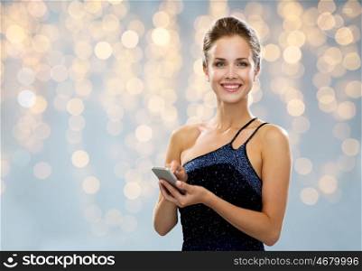 technology, smartphone, communication, people and holidays concept - smiling woman in evening dress holding smartphone over lights background