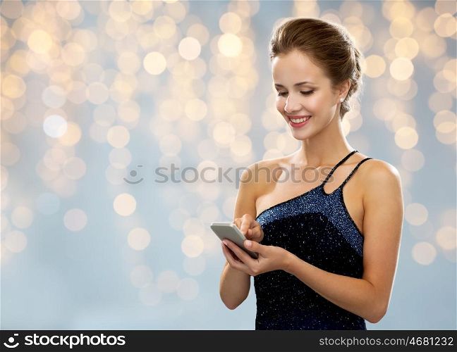 technology, smartphone, communication, people and holidays concept - smiling woman in evening dress texting on smartphone over lights background