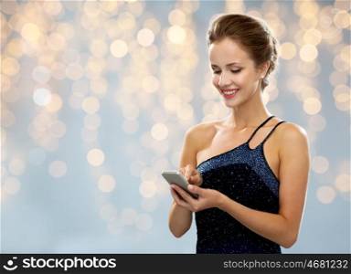 technology, smartphone, communication, people and holidays concept - smiling woman in evening dress texting on smartphone over lights background