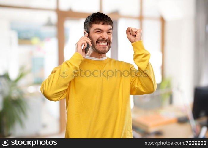 technology, success and people concept - happy smiling young man in yellow sweatshirt calling on smartphone over office background. happy smiling young man calling on smartphone