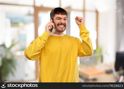technology, success and people concept - happy smiling young man in yellow sweatshirt calling on smartphone over office background. happy smiling young man calling on smartphone