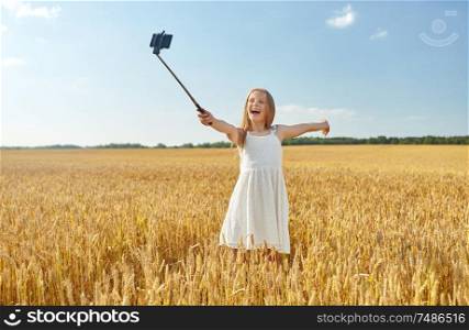 technology, summer and people concept - happy young girl in white dress taking picture by smartphone on selfie stick on cereal field. happy young girl taking selfie by smartphone