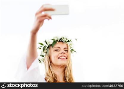 technology, summer holidays and people concept - smiling young woman in wreath of flowers taking selfie with smartphone