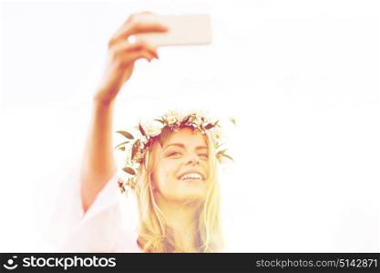 technology, summer holidays and people concept - smiling young woman in wreath of flowers taking selfie with smartphone. happy young woman taking smartphone selfie