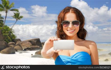 technology, summer holidays, travel and people concept - happy young woman in bikini swimsuit and sunglasses taking selfie with smatphone over beach and palm trees background. woman in swimsuit taking selfie with smatphone
