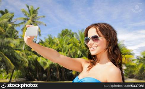 technology, summer holidays, travel and people concept - happy young woman in bikini swimsuit and sunglasses taking selfie with smatphone over sky and palm trees background. woman in swimsuit taking selfie with smatphone
