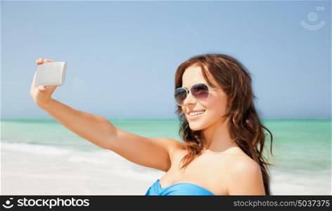 technology, summer holidays, travel and people concept - happy young woman in bikini swimsuit and sunglasses taking selfie with smatphone over beach background. woman in swimsuit taking selfie with smatphone