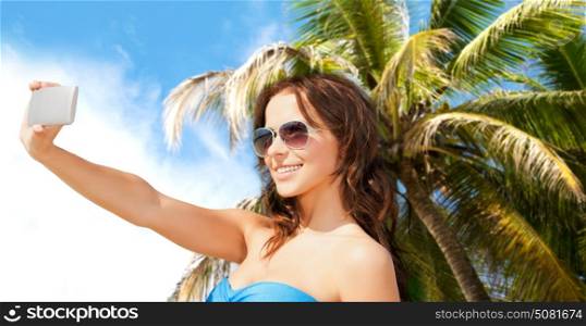technology, summer holidays, travel and people concept - happy young woman in bikini swimsuit and sunglasses taking selfie with smatphone over sky and palm trees background. woman in swimsuit taking selfie with smatphone