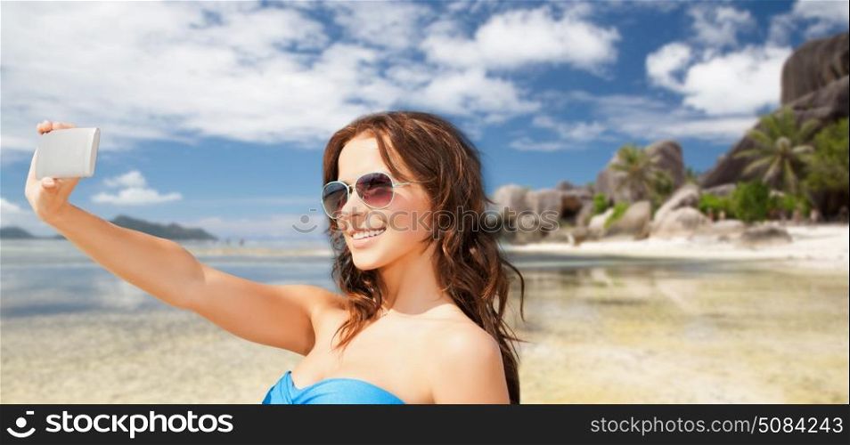 technology, summer holidays, travel and people concept - happy young woman in bikini swimsuit and sunglasses taking selfie with smatphone over beach and palm trees background. woman in swimsuit taking selfie with smatphone. woman in swimsuit taking selfie with smatphone