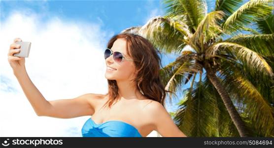 technology, summer holidays, travel and people concept - happy young woman in bikini swimsuit and sunglasses taking selfie with smatphone over sky and palm trees background. woman in swimsuit taking selfie with smatphone. woman in swimsuit taking selfie with smatphone