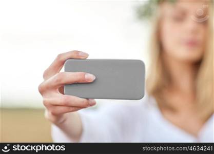 technology, summer holidays, vacation and people concept - close up of young woman in wreath of flowers taking picture by smartphone on cereal field