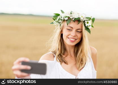 technology, summer holidays, vacation and people concept - smiling young woman in wreath of flowers taking selfie by smartphone on cereal field