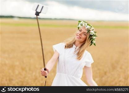 technology, summer holidays, vacation and people concept - smiling young woman in wreath of flowers taking picture by smartphone selfie stick on cereal field