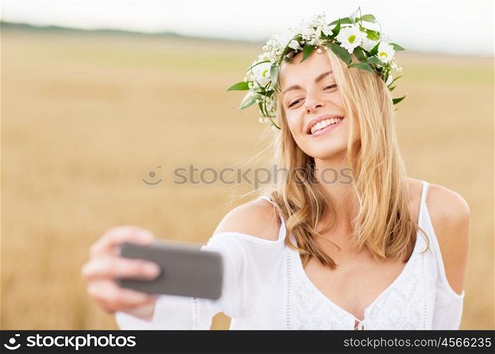 technology, summer holidays, vacation and people concept - smiling young woman in wreath of flowers taking selfie by smartphone on cereal field