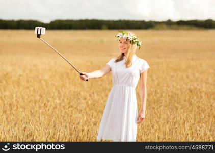 technology, summer holidays, vacation and people concept - smiling young woman in wreath of flowers taking picture by smartphone selfie stick on cereal field