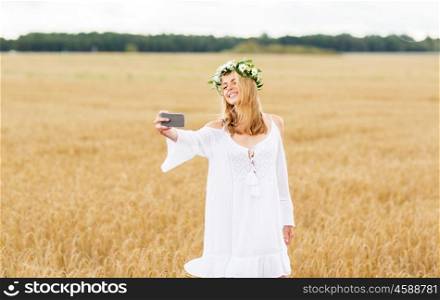 technology, summer holidays, vacation and people concept - smiling young woman in wreath of flowers taking selfie by smartphone on cereal field