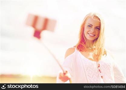 technology, summer holidays, vacation and people concept - smiling young woman in white dress taking picture by smartphone selfie stick on cereal field. happy young woman taking selfie by smartphone