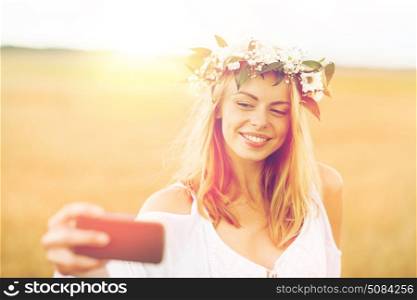 technology, summer holidays, vacation and people concept - smiling young woman in wreath of flowers taking selfie by smartphone on cereal field. happy young woman taking selfie by smartphone. happy young woman taking selfie by smartphone