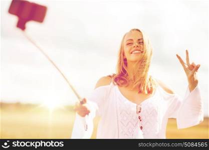 technology, summer holidays, vacation and people concept - smiling young woman in white dress taking picture by smartphone selfie stick and showing peace sign on cereal field. happy young woman taking selfie by smartphone. happy young woman taking selfie by smartphone