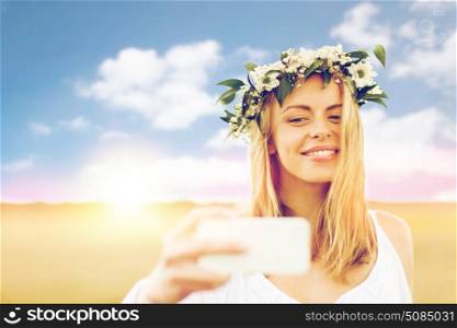 technology, summer holidays, vacation and people concept - smiling young woman in wreath of flowers taking selfie by smartphone on cereal field. happy young woman taking selfie by smartphone. happy young woman taking selfie by smartphone