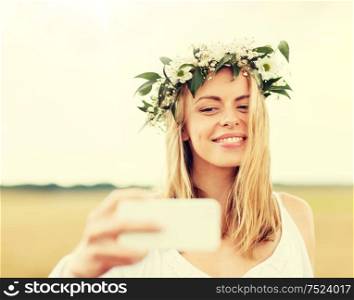 technology, summer holidays, vacation and people concept - smiling young woman in wreath of flowers taking selfie by smartphone on cereal field. happy young woman taking selfie by smartphone