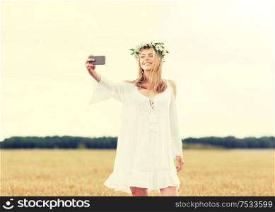 technology, summer holidays, vacation and people concept - smiling young woman in wreath of flowers taking selfie by smartphone on cereal field. happy young woman taking selfie by smartphone