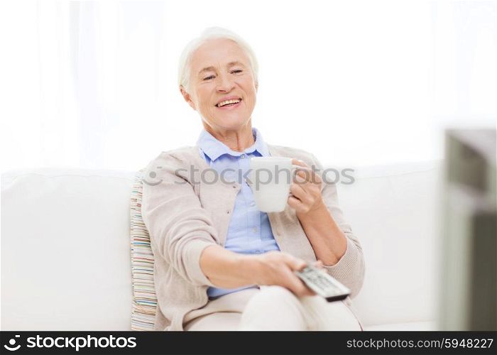 technology, television, age and people concept - happy senior woman watching tv, drinking tea and changing channels by remote control at home