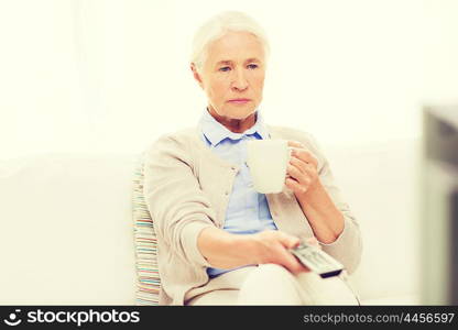technology, television, age and people concept - senior woman watching tv, drinking tea and changing channels by remote control at home
