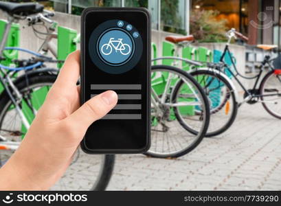 technology, transport and sustainability concept - close up of hand holding smartphone with mobile app and bicycle icon on screen over electric bike parking and charging station in city on background. hand with phone at electric bike charging station