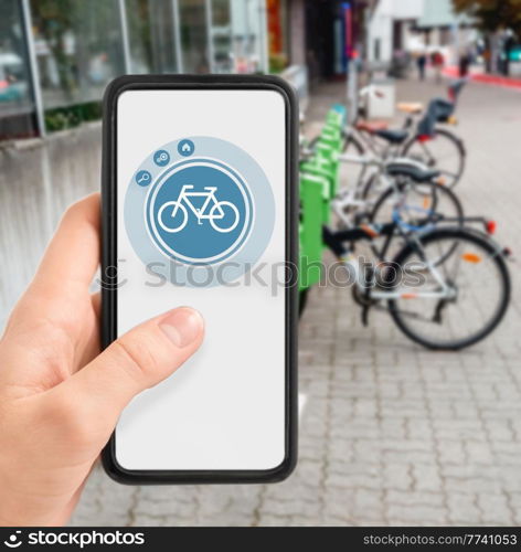 technology, transport and sustainability concept - close up of hand holding smartphone with mobile app and bicycle icon on screen over electric bike parking and charging station in city on background. hand with phone at electric bike charging station