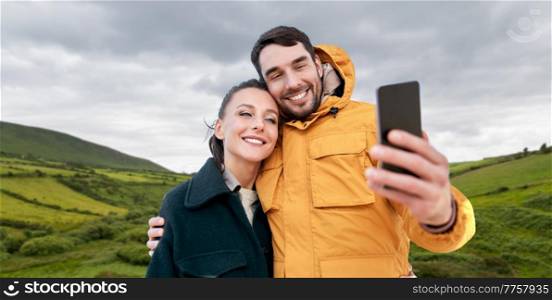technology, travel and tourism concept - happy couple taking selfie with smartphone over farmland fields and hills at wild atlantic way in ireland. couple taking selfie with smartphone in ireland